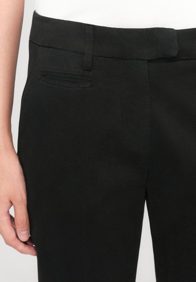 Pantalons noirs ajustés en tissu lisse, avec une taille légèrement texturée et un détail de poche cousu sur le côté droit.