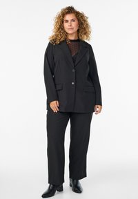 Black tailored blazer with two front pockets over a sheer polka dot top, paired with black straight-leg trousers and heeled ankle boots.