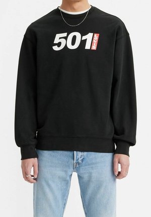 Osoba ubrana w czarną bluzę Levi's z białym logo "501" i czerwoną metką Levi's, w połączeniu z jasnoniebieskimi dżinsami i srebrnymi pierścionkami.