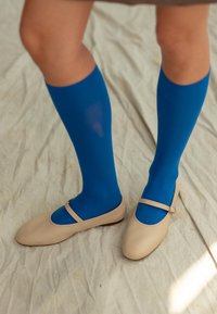 Bailarinas beige con una correa, combinadas con calcetas hasta la rodilla de un azul brillante. Los zapatos tienen una textura suave y mate y un diseño de punta redondeada.