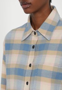 Chemise à boutons avec un motif à carreaux comprenant des couleurs bleues, beige et rose. Fabriquée en tissu doux avec un col pointu et des boutons noirs.