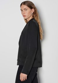 Schwarze Satinjacke mit lässiger Passform, langen Ärmeln und Seitentaschen. Glatte Textur und minimalistisches Design, ohne Muster oder Hardware.