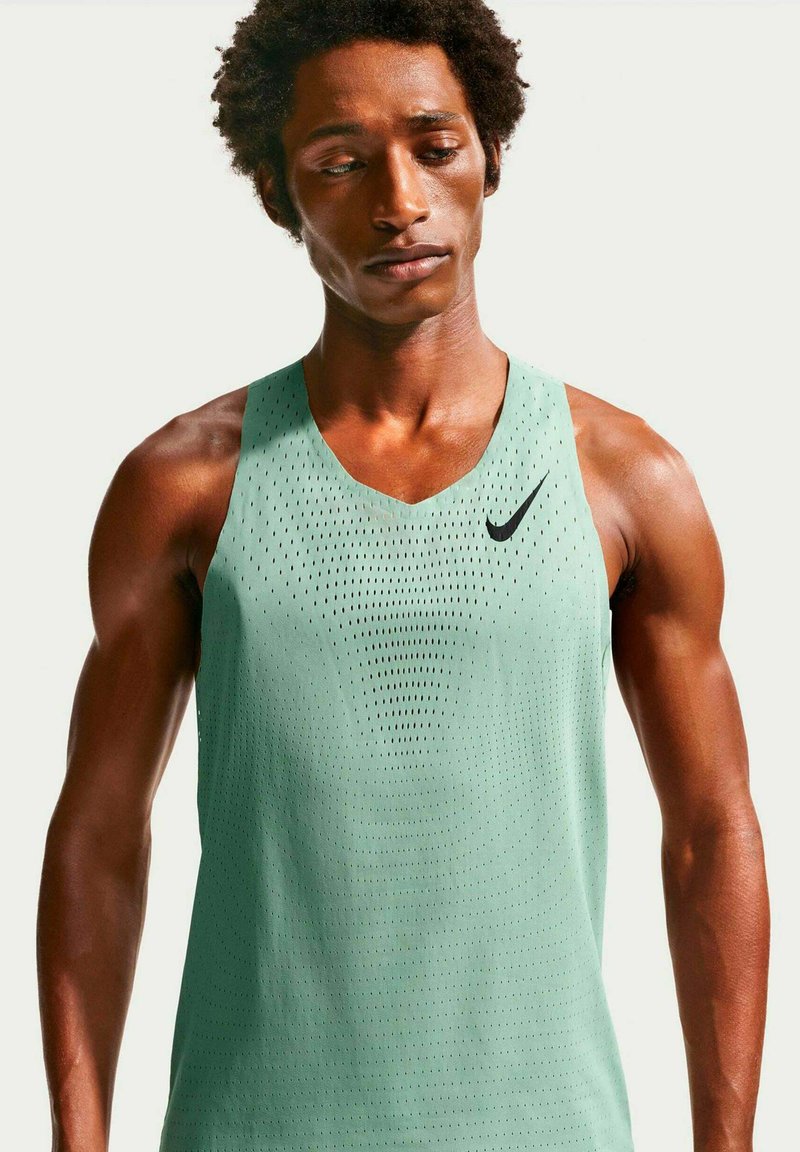 Homme portant un maillot de sport sans manches vert clair perforé Nike, regardant vers le bas, sur un fond clair uni.