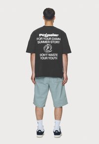 Černé oversized tričko s bílým textem na zádech, který zní: "Pegador PRO TVÝ PATRNÝ LETNÍ PŘÍBĚH NEZATRACUJ SVOU MLADOST." Kombinováno se světlými džíny šortkami.