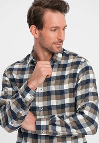 Camicia in flanella a quadri nei colori marrone, blu navy e bianco. Presenta un colletto button-down, maniche lunghe e una tasca sul petto. Texture morbida.
