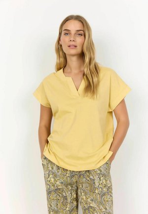Gelbes Kurzarmoberteil mit V-Ausschnitt, aus weichem Stoff. Kombiniert mit gemusterten Hosen im Paisleymuster in Gelb- und Blautönen.