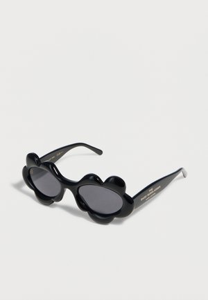 Czarne okulary przeciwsłoneczne z grubymi oprawkami w kształcie stokrotki i ciemnymi owalnymi szkłami, napis "The Daisy Sunglasses" na zauszniku.