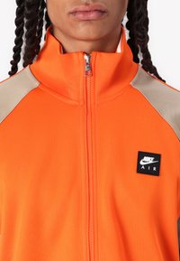 Narancssárga cipzáras sportdzseki bézs vállrész díszítéssel, kis fekete Nike Air logóval ellátva, sima, rugalmas anyagból készült.