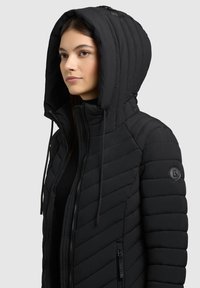 Black puffer jacket featuring a quilted design, high collar, and large hood. Includes zippered pockets and logo detail on the sleeve.