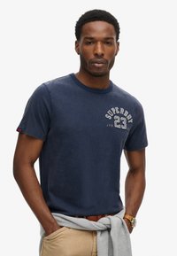 Dunkelblaues Baumwoll-T-Shirt mit Rundhalsausschnitt, präsentiert ein strukturiertes Logo-Print in Weiß und Grau auf der linken Brust. Lässiger Stil, kurze Ärmel.