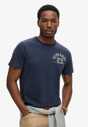 Navy blue cotton t-shirt with a crew neck, featuring a textured logo print in white and gray on the left chest. Casual style, short sleeves.