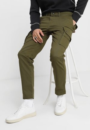 Persona con pantalones cargo verde oliva y zapatillas blancas sentada en un taburete blanco con una mano descansando sobre el muslo.