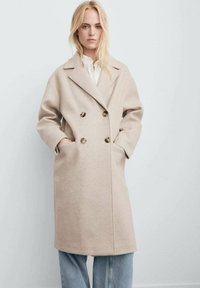 Manteau en laine beige avec un design à double boutonnage, grands revers et boutons dorés. Comprend des poches latérales et une coupe décontractée.