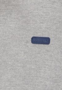 Lichtgrijze gebreide stof met een klein donkerblauw rechthoekig label waarop staat "J&JOY Every Step A New Journey" genaaid.