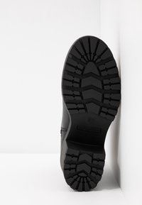 Semelle d'une botte noire avec des crampons profonds et dessinés, placée à la verticale contre un mur d'angle blanc et le sol.