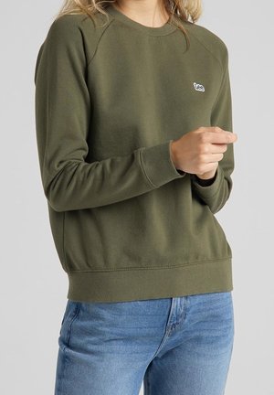 Osoba v olivově zelené mikině s logem "Lee" a modrých džínách, stojící před světlým šedým pozadím.