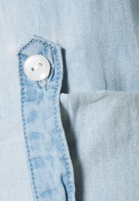 Ljusblå denimtyg med knappdetalj och ficka. Texturen är mjuk med en något blekt utseende och synlig sömnad.