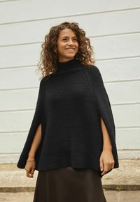Smiling woman with curly hair wearing a black knit poncho and brown skirt standing outdoors against a light wall background.