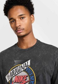 T-shirt nera in cotone con una texture sbiadita. Presenta un design grafico colorato con il testo "The Original Nike" ben in evidenza.