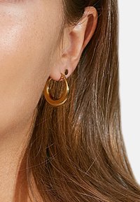 Kultaiset kehykset, joissa on sileä ja kiiltävä pinta, ja pienemmät kultaiset nappikorvakorut. Molemmissa on minimalistinen muotoilu ja hienovarainen tekstuuri.