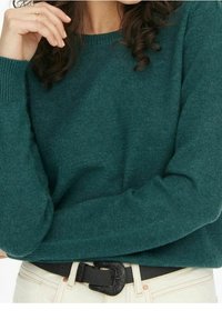 Maglione lavorato a maglia color teal con scollatura a girocollo, polsini a coste e una consistenza morbida, abbinato a una cintura nera con fibbia testurizzata su pantaloni chiari.