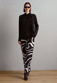 Zwarte button-up shirt met lange mouwen, gecombineerd met zwart-wit gestreepte broek. Model draagt zwarte zonnebril en houdt een zwarte tas vast.
