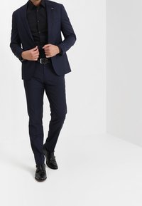 Marineblauwe pak met een op maat gemaakt jasje en slim-fit broek, gecombineerd met een zwart shirt en gepolijste zwarte schoenen. Gladde stofafwerking.