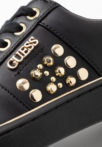 Primo piano di una sneaker nera con lacci neri, logo dorato "GUESS" e borchie decorative dorate sui lati sopra una suola spessa.
