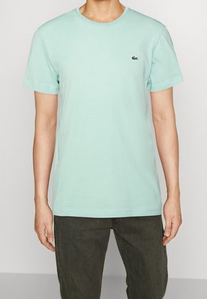 Mand iført mintgrøn kortærmet t-shirt med lille krokodillelogo på brystet og mørkegrå jeans, stående mod en ensfarvet baggrund.