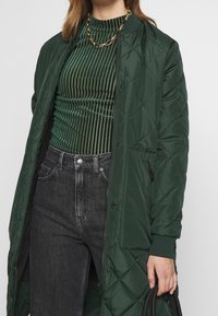 Grüne gesteppte Jacke mit Frontdruckknopfverschluss, getragen über einem grünen Rippentop, kombiniert mit schwarzen hochtaillierten Jeans. Schwarzes Accessoire in der Hand.