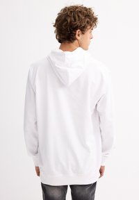 Sudadera blanca hecha de una tela suave. Presenta una capucha, mangas largas y un ajuste holgado. Combinada con jeans oscuros, fondo liso.