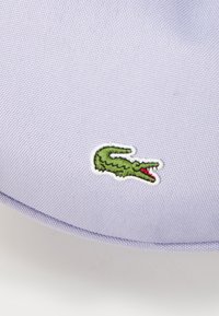 Sacca di tessuto viola con logo di coccodrillo ricamato in verde e accento rosso, texture liscia e forma arrotondata.