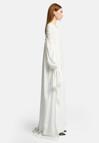 Langes weißes Kleid mit voluminösen, gerafften Ärmeln, hergestellt aus einem leichten Stoff. Die Silhouette ist fließend, mit einem bodenlangen Design.