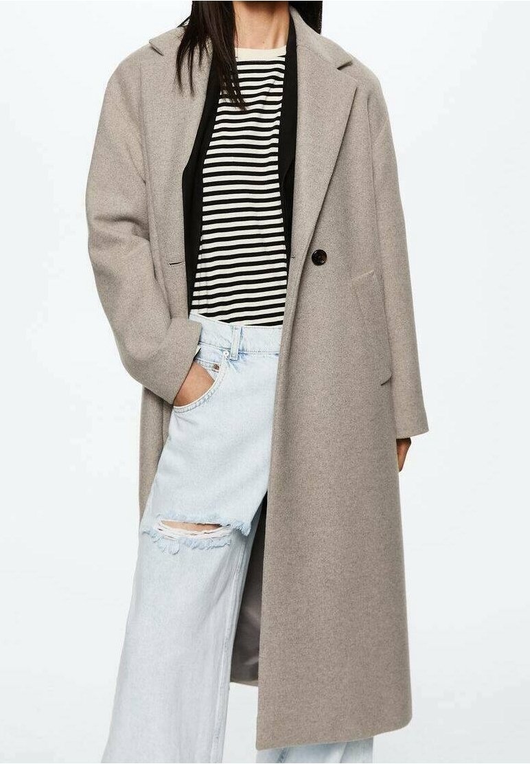 Femme portant un long manteau beige par-dessus une chemise rayée noire et blanche et un jean bleu clair déchiré, une main dans la poche.