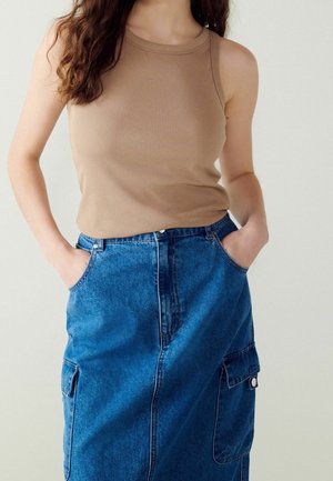 Kobieta w beżowym bezrękawnym prążkowanym topie włożonym do wysokiego jeansowego spódnicy z bocznymi kieszeniami naszywanymi, ręce w kieszeniach spódnicy.
