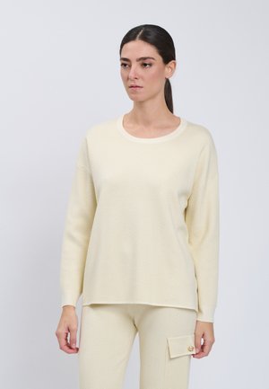 Frau mit dunklen Haaren in einem cremefarbenen Strickpullover und passenden Hosen mit Seitentasche und goldfarbenem Knopf, die vor einem schlichten weißen Hintergrund steht.