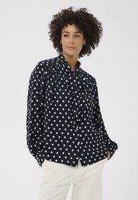 Blouse bleu marine à pois avec col noué, boutons sur le devant et coupe ample. Confectionnée en tissu lisse, elle présente des manches longues et des épaules froncées.