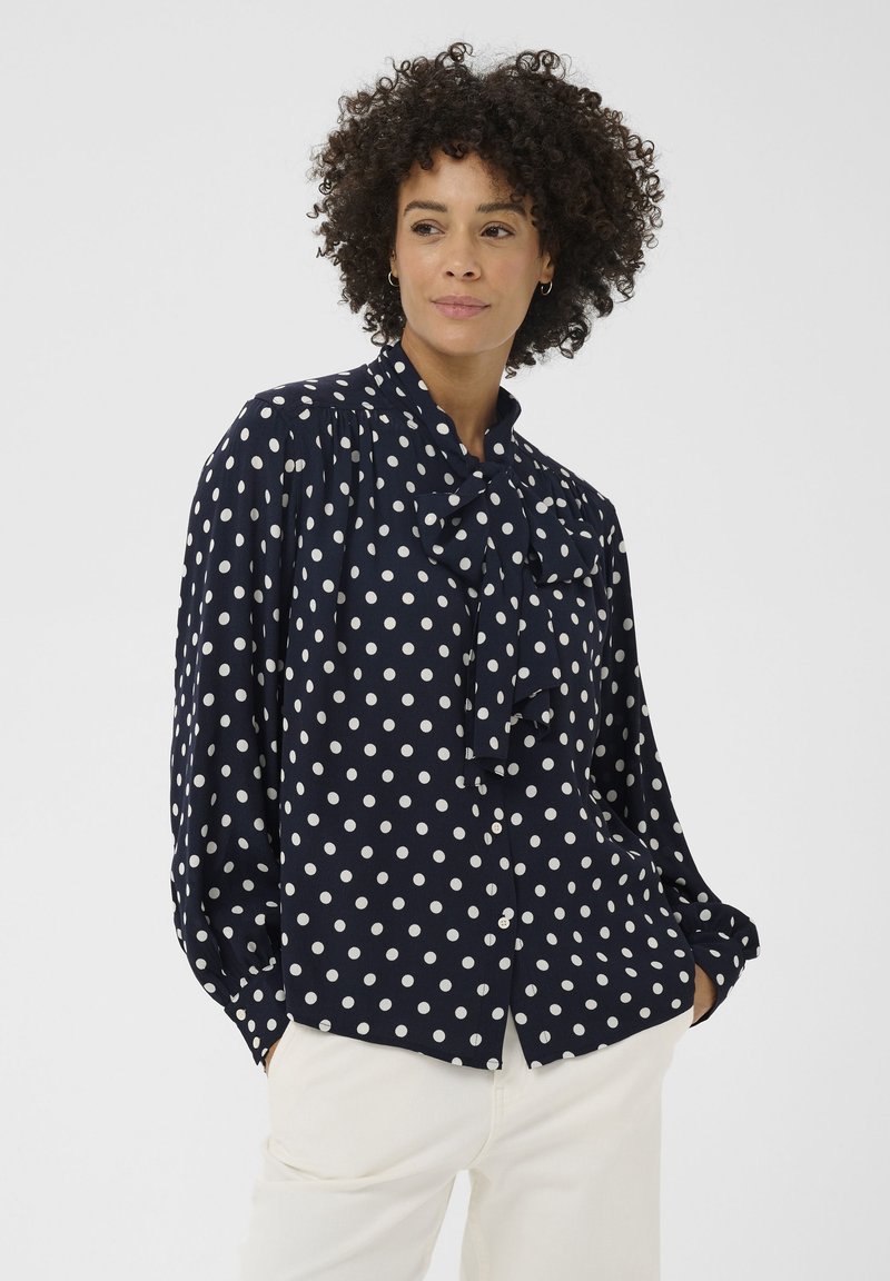 Blouse bleu marine à pois avec col noué, boutons sur le devant et coupe ample. Confectionnée en tissu lisse, elle présente des manches longues et des épaules froncées.