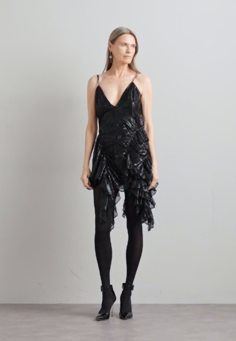 Robe noire froncée avec un décolleté en V profond, en tissu texturé. Portée avec des collants noirs et des bottines à bride de cheville. Boucles d'oreilles simples comme accents.