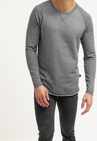 Felpa grigia a maniche lunghe con texture a costine, maniche raglan e orlo arrotondato. Presenta cuciture sottili e un'etichetta laterale.