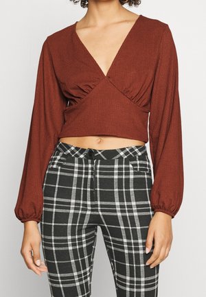 Žena v hnědém dlouhém rukávu s krátkým topem a výstřihem do V, s černobílými kostkovanými kalhotami s vysokým pasem, stojící s uvolněnýma rukama.