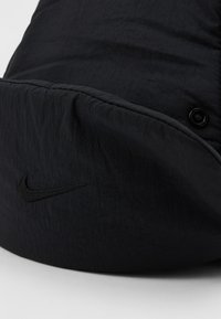 Melna Nike cepure ar izšūtu melnu swoosh logotipu un aizmugurē esošu spiedpogu uz fona ar vienkāršu gaišu krāsu.