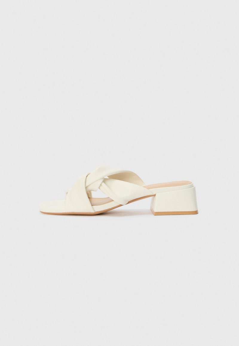 Sandales à talons bloc couleur crème avec des brides torsadées et un lit de pied plat. Matériau synthétique lisse avec une finition mate.