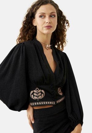 Mujer con cabello rizado que lleva una blusa negra con mangas voluminosas y una cintura bordada, combinada con una falda negra y un collar con colgante.