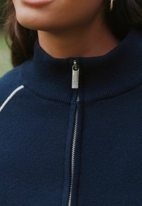 Pull marine à fermeture éclair avec col montant ; présente une fermeture éclair argentée et une texture côtelée. Coutures blanches remarquables le long des épaules.