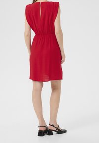 Robe courte rouge avec une taille cintrée, de courtes manches gigot et une fermeture au dos en forme de goutte. Associée à des sandales à talons noires. Texture de tissu lisse.