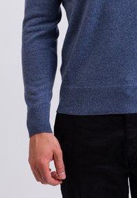 Blauer Wollpullover mit gerippten Bündchen und Saum, der eine glatte Textur aufweist. Eine Hand liegt nahe dem unteren Ende des Kleidungsstücks.