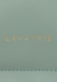 Pelle testurizzata verde chiaro con logo "EXPATRIÉ" in oro a rilievo. Presenta linee pulite e un sottile dettaglio di cucitura lungo i bordi.