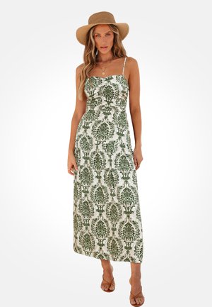 Kobieta ubrana w białą, bezrękawnikową sukienkę maxi z zielonym wzorem damasceńskim, szerokim kapeluszem w kolorze jasno-brązowym, nałożonymi warstwowo złotymi naszyjnikami i brązowymi sandałami.