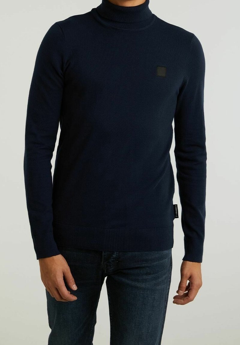 Marinblå turtleneck-tröja i ribbad textur, gjord av mjukt material. Har en liten svart lappar på bröstet och åtsittande ärmslut.